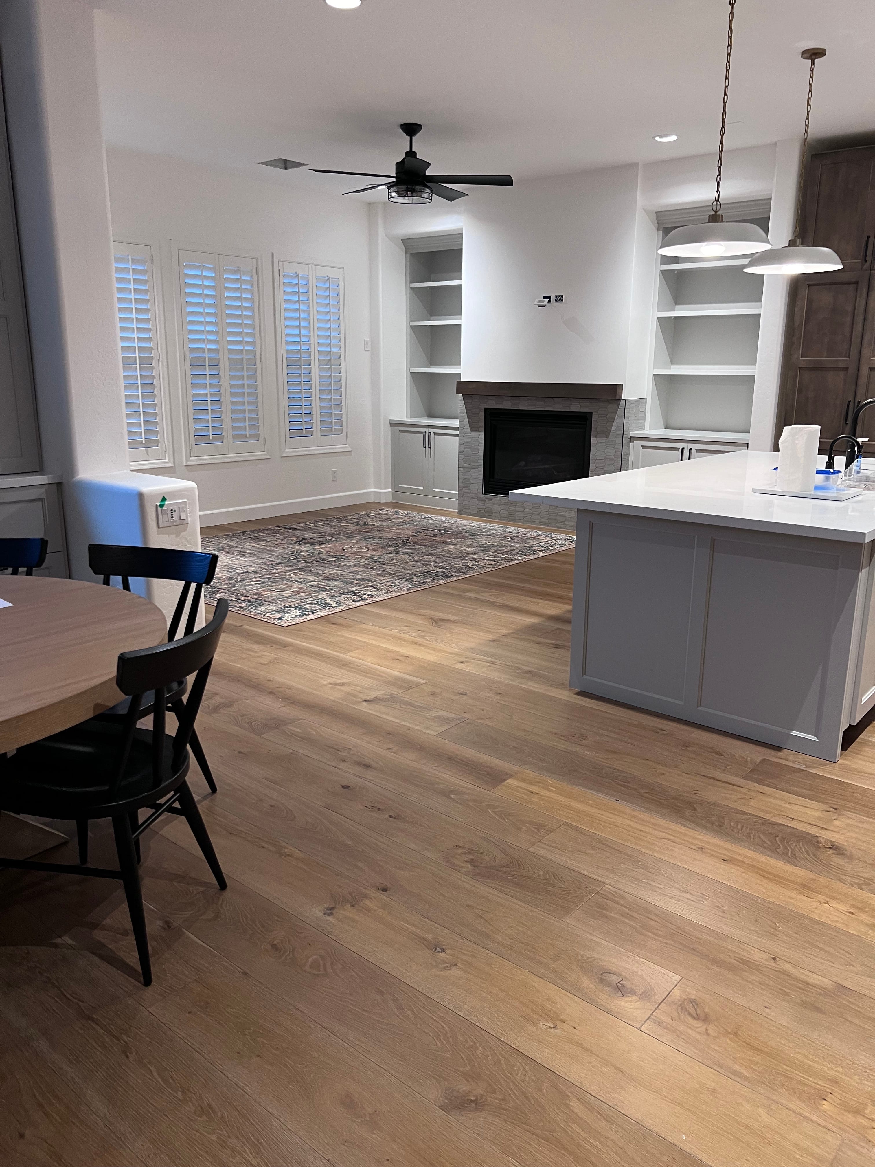 Modern open-concept living and dining room with light wood floors, a gray kitchen island, a fireplace flanked by built-in shelves, a dining table, and windows with white shutters.